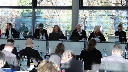 Mehrere Personen sitzen hinter einem langen Tisch vor großen Fenstern mit Blick ins Freie, arbeiten an Laptops oder hören zu, während davor weitere Teilnehmende Platz genommen haben, wodurch das Bild eine Gremiensitzung oder Fachdebatte dokumentiert und für strukturierte Zusammenarbeit und gemeinsame Entscheidungsfindung steht.