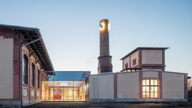 Sanierter Industriebau mit gläsernem Eingangsbereich und historischem Schornstein in der Abenddämmerung.