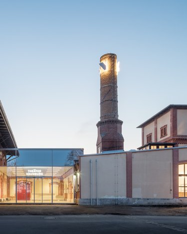 Sanierter Industriebau mit gläsernem Eingangsbereich und historischem Schornstein in der Abenddämmerung.