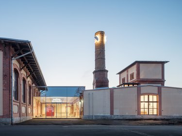 Sanierter Industriebau mit gläsernem Eingangsbereich und historischem Schornstein in der Abenddämmerung.