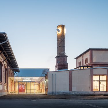 Sanierter Industriebau mit gläsernem Eingangsbereich und historischem Schornstein in der Abenddämmerung.