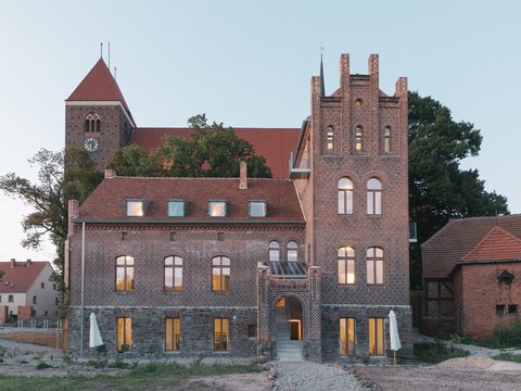 „Außenansicht eines historischen Backsteingebäudes mit Zinnenturm und beleuchteten Fenstern in der Dämmerung, im Hintergrund ein Kirchturm.“