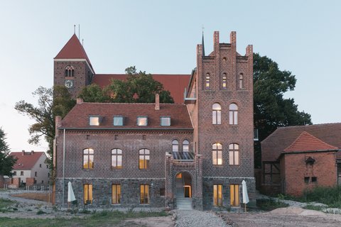 „Außenansicht eines historischen Backsteingebäudes mit Zinnenturm und beleuchteten Fenstern in der Dämmerung, im Hintergrund ein Kirchturm.“