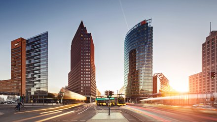 Stadtansicht: Breiter Platz bei Sonnenuntergang mit drei markanten Hochhäusern; gelber Doppeldeckerbus und Lichtspuren des Verkehrs; Schilder „DB“ und „Bahnhof Potsdamer Platz“.