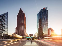 Stadtansicht: Breiter Platz bei Sonnenuntergang mit drei markanten Hochhäusern; gelber Doppeldeckerbus und Lichtspuren des Verkehrs; Schilder „DB“ und „Bahnhof Potsdamer Platz“.