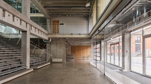 Heller mehrstöckiger Innenraum mit Tribüne auf der linken Seite und einer Fensterfront auf der rechten Seite.