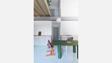 Innenraum mit grünem Tisch Alt: Küchen‑/Essbereich mit hohem Luftraum; grüner Holztisch, orange Kinderstuhl, weiße Küchenzeile; oben Holzträger und Maschendrahtbrüstung; runde Pendelleuchte.