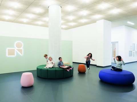 "Moderner heller Kinderbereich mit weißer Decke und indirekter Beleuchtung. Zentral ein weißer Pfeiler. Wände teilweise in zartem Grün und Weiß. Mehrere Kinder spielen verteilt im Raum: auf einem grünen runden Sitzelement in der Mitte, auf einem rosa Sitzsack vorne links, auf einem orangen Sitzsack rechts und in der Mitte. Ein weißes Neon-Logo an der grünen Wand. Blaugrauer Bodenbelag."