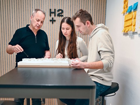 "Drei Personen arbeiten gemeinsam an einem weißen Architekturmodell an einem schwarzen Tisch. Links steht ein älterer Mann in schwarzem Poloshirt, der auf das Modell zeigt. In der Mitte sitzt eine junge Frau mit langen braunen Haaren in hellgrauem Pullover. Rechts sitzt ein junger Mann in hellbeigem Hoodie in Jeans. Alle drei konzentrieren sich auf das Modell. Im Hintergrund ist eine Wand mit vertikalen Holzlatten in Natur- und Grautönen sichtbar, darauf ein großes „h2+" Logo in schwarzen Buchstaben. Auf der rechten Seite an der Wand hängt eine Kunstinstallation aus blauen und gelben geometrischen Formen. Der Boden ist mit Holzparkett belegt."