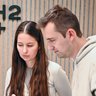 "Drei Personen arbeiten gemeinsam an einem weißen Architekturmodell an einem schwarzen Tisch. Links steht ein älterer Mann in schwarzem Poloshirt, der auf das Modell zeigt. In der Mitte sitzt eine junge Frau mit langen braunen Haaren in hellgrauem Pullover. Rechts sitzt ein junger Mann in hellbeigem Hoodie in Jeans. Alle drei konzentrieren sich auf das Modell. Im Hintergrund ist eine Wand mit vertikalen Holzlatten in Natur- und Grautönen sichtbar, darauf ein großes „h2+" Logo in schwarzen Buchstaben. Auf der rechten Seite an der Wand hängt eine Kunstinstallation aus blauen und gelben geometrischen Formen. Der Boden ist mit Holzparkett belegt."