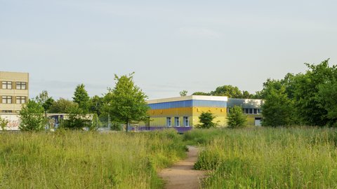  Eine Weitwinkelaufnahme eines modernen, flachen Gebäudes, das hinter einer hohen Wiese mit einem Feldweg steht. Das Gebäude hat eine farbenfrohe Fassade mit horizontalen Streifen in Lila, Gelb und Blau. Links davon steht ein älteres, mehrstöckiges Betongebäude. Bäume und ein klarer Himmel bilden den Hintergrund.
