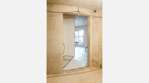 Rohbau-Innenraum mit rund ausgeschnittener Betonöffnung; dahinter weißer Trennwandflur und ein Raum mit gebogener Außenwand und Fenster; freiliegende Kabel an Decke und Wänden, unfertiger Boden.