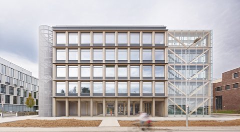 „Frontansicht eines modernen fünfstöckigen Bürogebäudes mit markanter Holzrasterfassade, einem gläsernen Treppenhausanbau und einem zylindrischen Metallturm.“