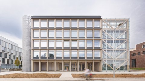 „Frontansicht eines modernen fünfstöckigen Bürogebäudes mit markanter Holzrasterfassade, einem gläsernen Treppenhausanbau und einem zylindrischen Metallturm.“