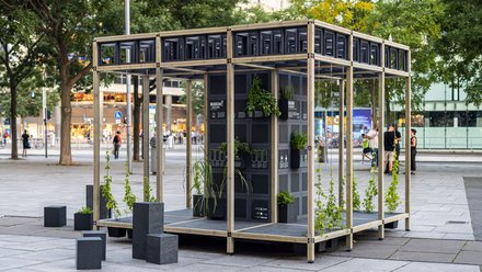 Offener Holzpavillon im Stadtraum mit Infotafeln und integrierter Begrünung.