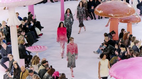 "Modenschau in einem hellen Raum mit fantasievoller Bühneninstallation aus übergroßen Pilzskulpturen in Rosa- und Rottönen. Mehrere Models laufen über den Laufsteg, umgeben von einem sitzenden Publikum. Die Kleidung ist farbenreich und gemustert, während viele Zuschauer die Szene mit Smartphones fotografieren."