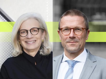 Zwei Personen in formeller Kleidung stehen nebeneinander vor einer modernen Gebäudefassade