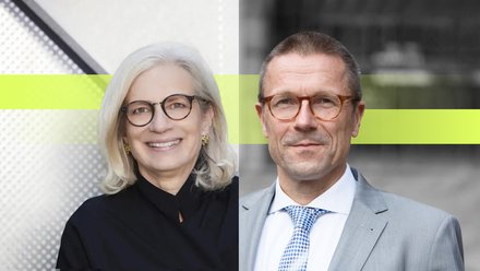 Zwei Personen in formeller Kleidung stehen nebeneinander vor einer modernen Gebäudefassade