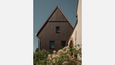 Nahaufnahme der Giebelseite eines zweistöckigen Hauses mit dunkelbrauner Holzverkleidung und einem steilen roten Ziegeldach. Das Haus hat drei schwarze quadratische Fenster, eines auf der Giebelebene und zwei darunter. Links vom Haus ist ein silbernes Regenfallrohr zu sehen. Rechts führt eine weiße verputzte Wand mit einem ockerfarbenen Rundbogen in den Hintergrund. Im Vordergrund, unter dem Haus, blühen rosa und weiße Rosensträucher. Der Himmel ist klar und blau.