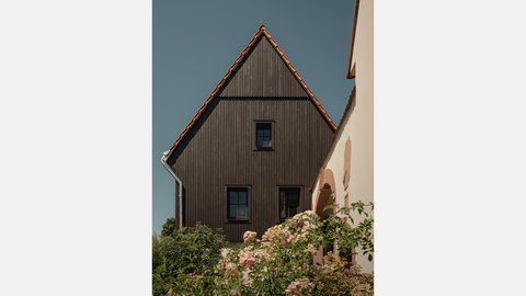 Nahaufnahme der Giebelseite eines zweistöckigen Hauses mit dunkelbrauner Holzverkleidung und einem steilen roten Ziegeldach. Das Haus hat drei schwarze quadratische Fenster, eines auf der Giebelebene und zwei darunter. Links vom Haus ist ein silbernes Regenfallrohr zu sehen. Rechts führt eine weiße verputzte Wand mit einem ockerfarbenen Rundbogen in den Hintergrund. Im Vordergrund, unter dem Haus, blühen rosa und weiße Rosensträucher. Der Himmel ist klar und blau.