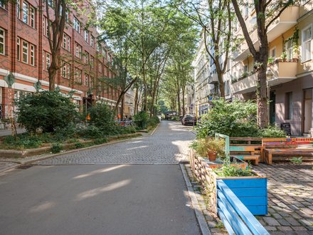 Ruhige, baumbestandene Wohnstraße mit Sitzbänken und Pflanzkästen am Straßenrand.