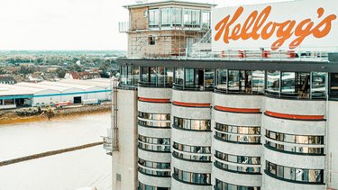 Mehrstöckiges Gebäude mit abgerundeten Fenstern und Schriftzug 'Kellogg's' auf dem Dach, daneben ein modernes Gebäude mit Glasfassade.
