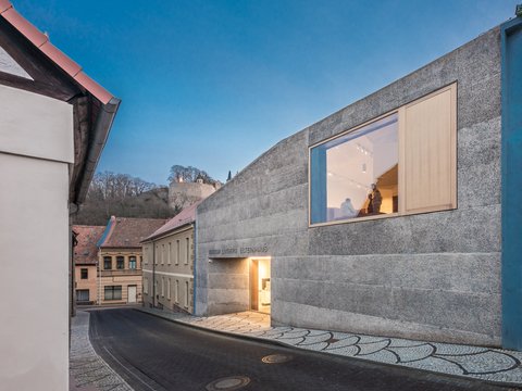 "Modernes Museumsgebäude mit grauer Betonfassade und blau lackierter Metallpfoste an einer engen Gasse. Ein großes horizontales Fenster mit hellem Holzrahmen zeigt den beleuchteten Innenraum mit zwei Besuchern. Darunter ein gelb beleuchteter Eingang. Links sind historische Häuser mit gelblicher und rosa Fassade sichtbar. Der Boden ist gepflastert mit geometrischen Mustern. Der Himmel ist klar und blau."