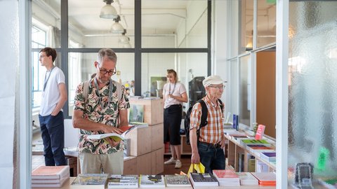 „Blick durch eine Glasscheibe in einen hellen Raum, in dem mehrere Personen an einer Auslage mit Büchern und Broschüren stöbern.“