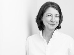 "Schwarz-Weiß-Porträtfoto einer Frau mittleren Alters mit schulterlangem, welligem dunklem Haar und Seitenscheitel. Sie trägt ein weißes Hemd mit V-Ausschnitt und blickt ruhig zur Seite in die Kamera. Sie hat einen feinen, subtilen Gesichtsausdruck. Der Hintergrund ist neutral und hell. Das Foto hat einen professionellen, zeitlosen Charakter."