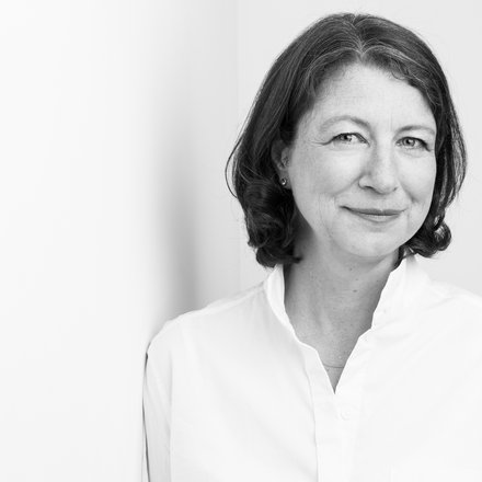 "Schwarz-Weiß-Porträtfoto einer Frau mittleren Alters mit schulterlangem, welligem dunklem Haar und Seitenscheitel. Sie trägt ein weißes Hemd mit V-Ausschnitt und blickt ruhig zur Seite in die Kamera. Sie hat einen feinen, subtilen Gesichtsausdruck. Der Hintergrund ist neutral und hell. Das Foto hat einen professionellen, zeitlosen Charakter."