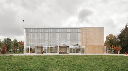Lange Gebäudeansicht: zweigeschossige Glasfront mit Vorhängen links, geschlossene Holzverkleidung rechts; Wiese davor.