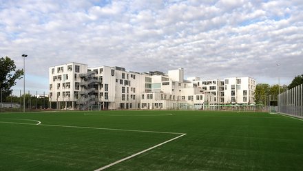 Großer Sportplatz vor ausgedehntem weißem Schulkomplex mit außenliegenden Treppen; bewölkter Himmel.