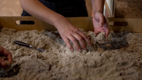 "Nahaufnahme von Händen, die feinen, hellen Sand in einer flachen Holzkiste formen. Ein kleines Werkzeug liegt daneben. Die Finger greifen und modellieren den Sand, wodurch kleine Hügel entstehen. Die Szene zeigt eine haptische, interaktive Ausstellungssituation."