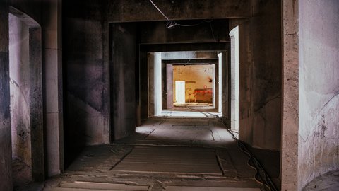 Langer, dunkler Betongang mit hintereinanderliegenden rechteckigen Durchgängen; staubiger Boden, Kabel rechts entlang und Leitung von der Decke; am Ende ein warm beleuchteter Raum mit rotem Element an der Wand.