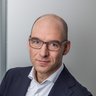 "Porträt eines Mannes mittleren Alters mit Brille und dunklem Blazer vor neutralem grauem Hintergrund. Er trägt ein weißes Hemd und blickt direkt in die Kamera."