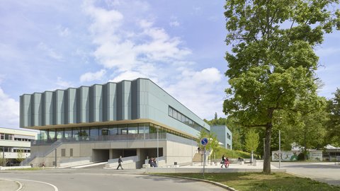 Moderner mehrstöckiger Gebäudekomplex mit großen Fenstern und umlaufendem Vordach neben einer Straße und einem Baum unter blauem Himmel