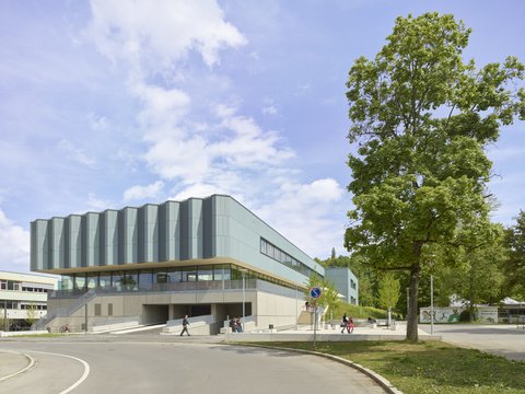 Moderner mehrstöckiger Gebäudekomplex mit großen Fenstern und umlaufendem Vordach neben einer Straße und einem Baum unter blauem Himmel
