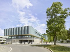 Moderner mehrstöckiger Gebäudekomplex mit großen Fenstern und umlaufendem Vordach neben einer Straße und einem Baum unter blauem Himmel