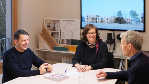 Drei Personen sitzen in einem Architekturbüro an einem Tisch vor Modellen und Bauplänen.