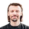 "Porträt eines Mannes mit schulterlangen braunen Haaren und Bartansatz. Er trägt ein schwarzes Sweatshirt und eine Smartwatch. Seine Arme sind verschränkt vor dem Körper, er schaut direkt in die Kamera und lächelt leicht. Der Hintergrund ist weiß."