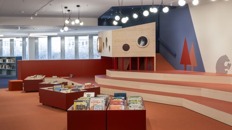 "Farbenfroher Kinderbibliotheksbereich. Wände mit gelben Akzenten und dunkelblauen Paneelen. Rechts großes gelbes Wandregal mit Bücherausstellung und "Lesecafé"-Schriftzug. Zentral ein kreatives Raumdesign mit: roten Sitzbänken als Regalelemente mit Büchern, dahinter ein Holzhaus-ähnliches Element mit Rundöffnungen und roten Bäumen-Grafikelementen. Blaue und orange Teppichzonen. Mehrere Sessel in Gelb und Anthrazit. Dunkelblaue Fensterfront."
