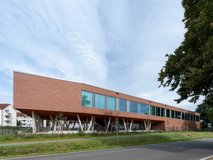 "Modernes Gebäude mit roter Backsteinfassade und großzügiger Glasfront. Das langgestreckte, einstöckige Bauwerk steht auf weißen Betonstützen, die einen offenen Untergeschoss-Bereich schaffen. Eine Reihe großflächiger Fenster erstreckt sich über die gesamte Länge der Fassade. Rechts im Bild ein dichter grüner Baum. Im Vordergrund grüne Rasenflächen und eine asphaltierte Zufahrt mit Leitpfosten. Im Hintergrund links sind Wohngebäude sichtbar. Der Himmel ist teilweise bewölkt und hell."