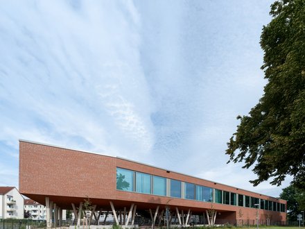 "Modernes Gebäude mit roter Backsteinfassade und großzügiger Glasfront. Das langgestreckte, einstöckige Bauwerk steht auf weißen Betonstützen, die einen offenen Untergeschoss-Bereich schaffen. Eine Reihe großflächiger Fenster erstreckt sich über die gesamte Länge der Fassade. Rechts im Bild ein dichter grüner Baum. Im Vordergrund grüne Rasenflächen und eine asphaltierte Zufahrt mit Leitpfosten. Im Hintergrund links sind Wohngebäude sichtbar. Der Himmel ist teilweise bewölkt und hell."