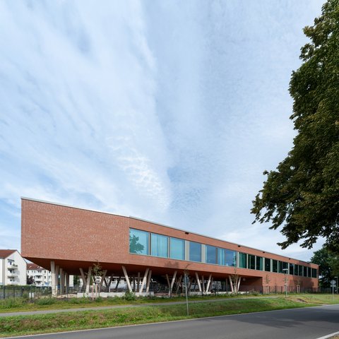 "Modernes Gebäude mit roter Backsteinfassade und großzügiger Glasfront. Das langgestreckte, einstöckige Bauwerk steht auf weißen Betonstützen, die einen offenen Untergeschoss-Bereich schaffen. Eine Reihe großflächiger Fenster erstreckt sich über die gesamte Länge der Fassade. Rechts im Bild ein dichter grüner Baum. Im Vordergrund grüne Rasenflächen und eine asphaltierte Zufahrt mit Leitpfosten. Im Hintergrund links sind Wohngebäude sichtbar. Der Himmel ist teilweise bewölkt und hell."