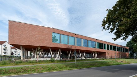 "Modernes Gebäude mit roter Backsteinfassade und großzügiger Glasfront. Das langgestreckte, einstöckige Bauwerk steht auf weißen Betonstützen, die einen offenen Untergeschoss-Bereich schaffen. Eine Reihe großflächiger Fenster erstreckt sich über die gesamte Länge der Fassade. Rechts im Bild ein dichter grüner Baum. Im Vordergrund grüne Rasenflächen und eine asphaltierte Zufahrt mit Leitpfosten. Im Hintergrund links sind Wohngebäude sichtbar. Der Himmel ist teilweise bewölkt und hell."