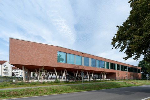 "Modernes Gebäude mit roter Backsteinfassade und großzügiger Glasfront. Das langgestreckte, einstöckige Bauwerk steht auf weißen Betonstützen, die einen offenen Untergeschoss-Bereich schaffen. Eine Reihe großflächiger Fenster erstreckt sich über die gesamte Länge der Fassade. Rechts im Bild ein dichter grüner Baum. Im Vordergrund grüne Rasenflächen und eine asphaltierte Zufahrt mit Leitpfosten. Im Hintergrund links sind Wohngebäude sichtbar. Der Himmel ist teilweise bewölkt und hell."
