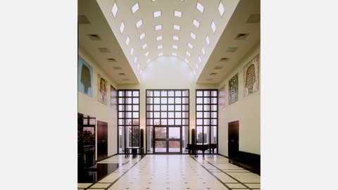 "Moderner Ausstellungsraum mit hochgewölbter Decke und rautenförmigen Oberlichtern in zwei Reihen. Creme-farbene Wände, große dreiteilige Fensterfront mit Ausblick auf herbstliche Vegetation. Vier dekorative Kunstobjekte an den Seitenwänden. Glatte Bodenfliesen mit Mosaikmuster. Zwei schwarze Stühle im Raum."