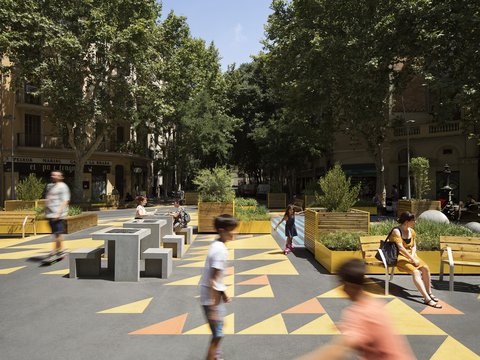 "Lebendiger urbaner Platz mit geometrischem Muster aus gelben, orange und roten Farbflächen. Mehrere Menschen verschiedenen Alters bewegen sich über den Platz – einige spazieren, andere sitzen auf hellen Sitzelementen. Links und rechts säumen dichte grüne Bäume den Platz. Historische Wohn- und Geschäftshäuser mit warmem Sandsteinton rahmen die Szene ein. Im Hintergrund sind weitere Bäume erkennbar."