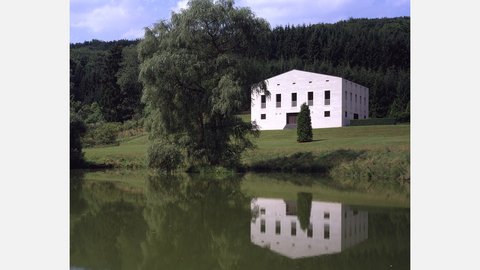 "Weißes quadratisches Betongebäude mit flachem Dach in ländlicher Waldlandschaft. Große rechteckige Fensteröffnungen auf der sichtbaren Seite. Umgeben von Wiesen und dichten grünen Wald. Klarer Himmel mit Wolken."