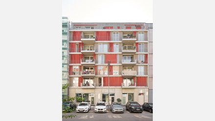 Mehrgeschossiges Wohnhaus mit Balkonen und roten Vorhängen über einem Parkplatz.